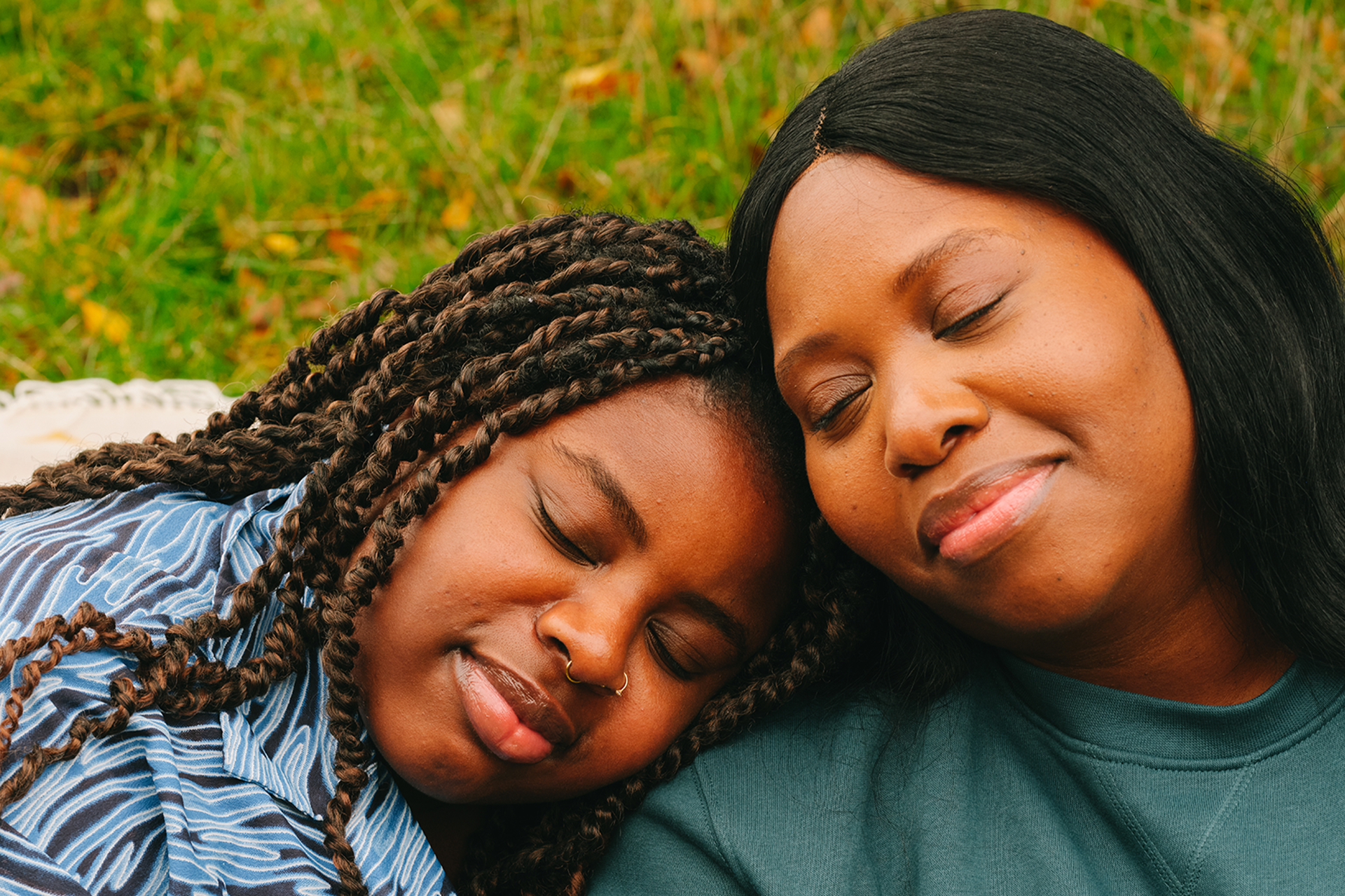 A young Black woman and an older Black woman leaning their heads on each other's shoulders and smiling. Their eyes are closed.