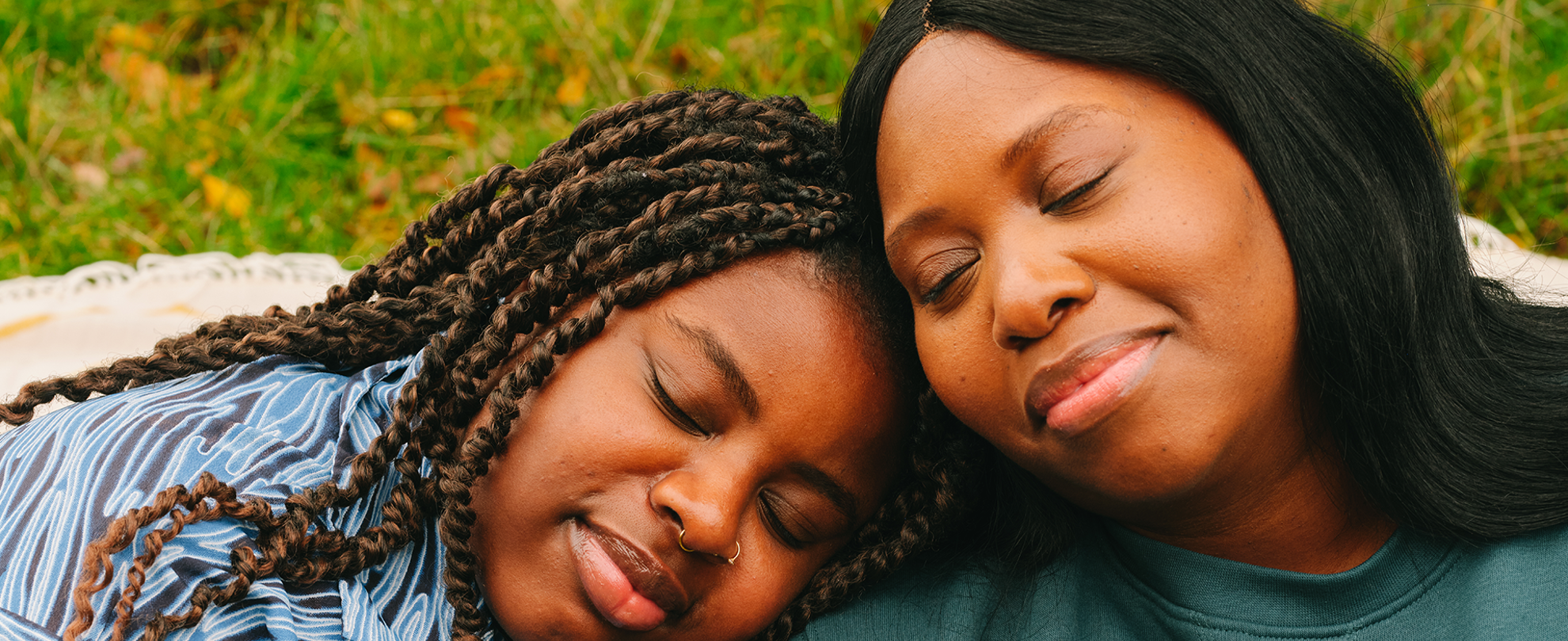 A young Black woman and an older Black woman leaning their heads on each other's shoulders and smiling. Their eyes are closed.