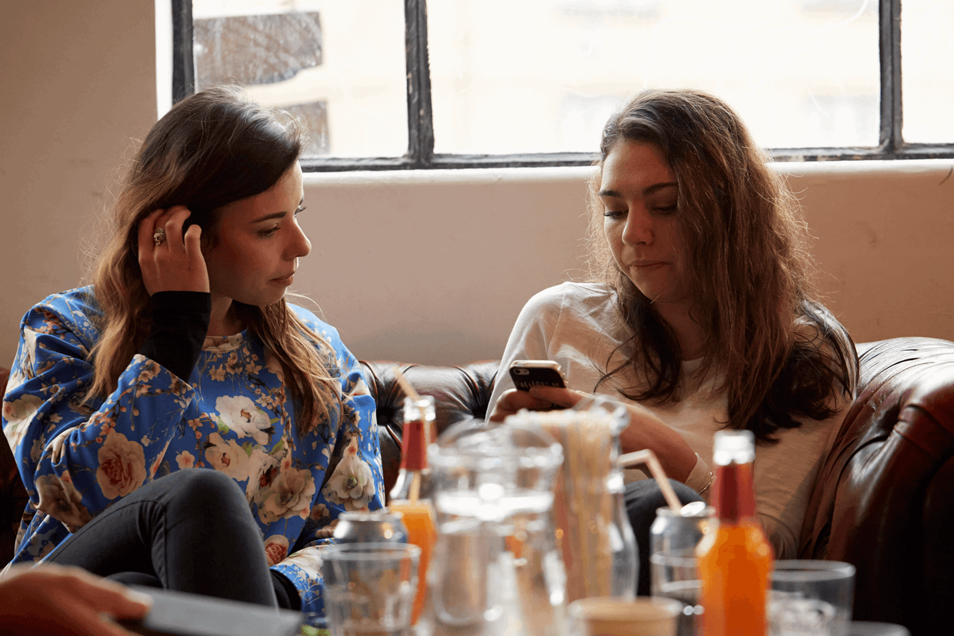 medium shot of two girls sitting on a couch one is using a phone while another girl is glancing on her phone bottles and drinks are on the table as foreground