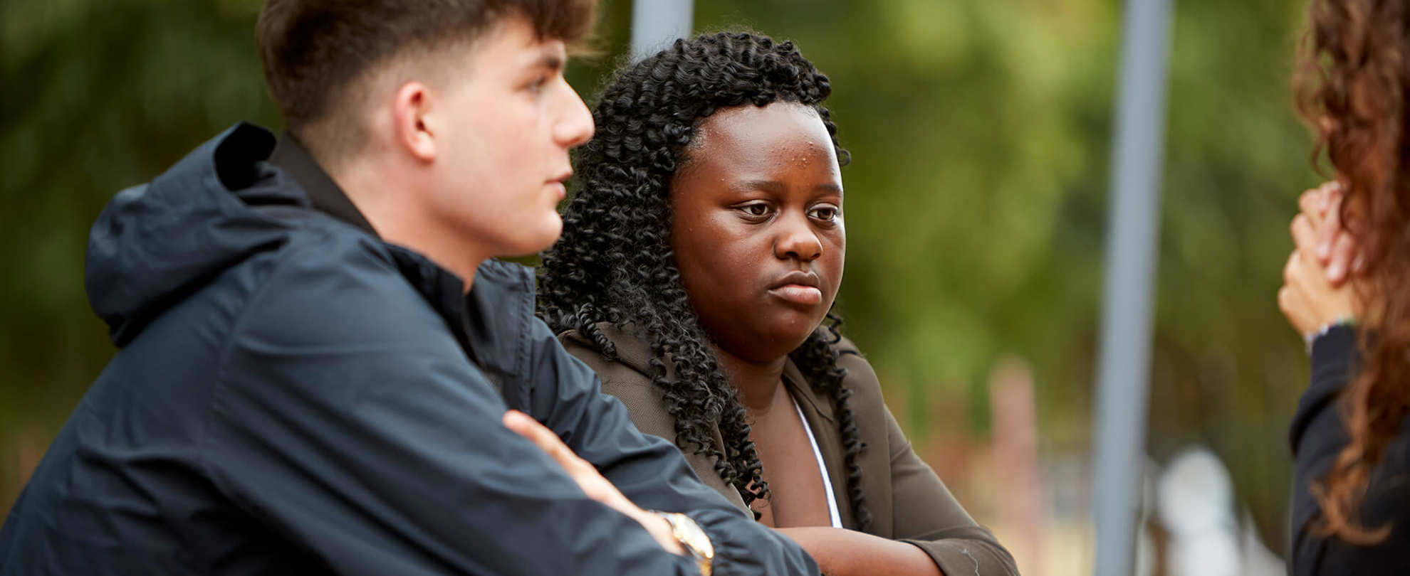 Two young people listening intently to their friend while sitting on a bench in a park.