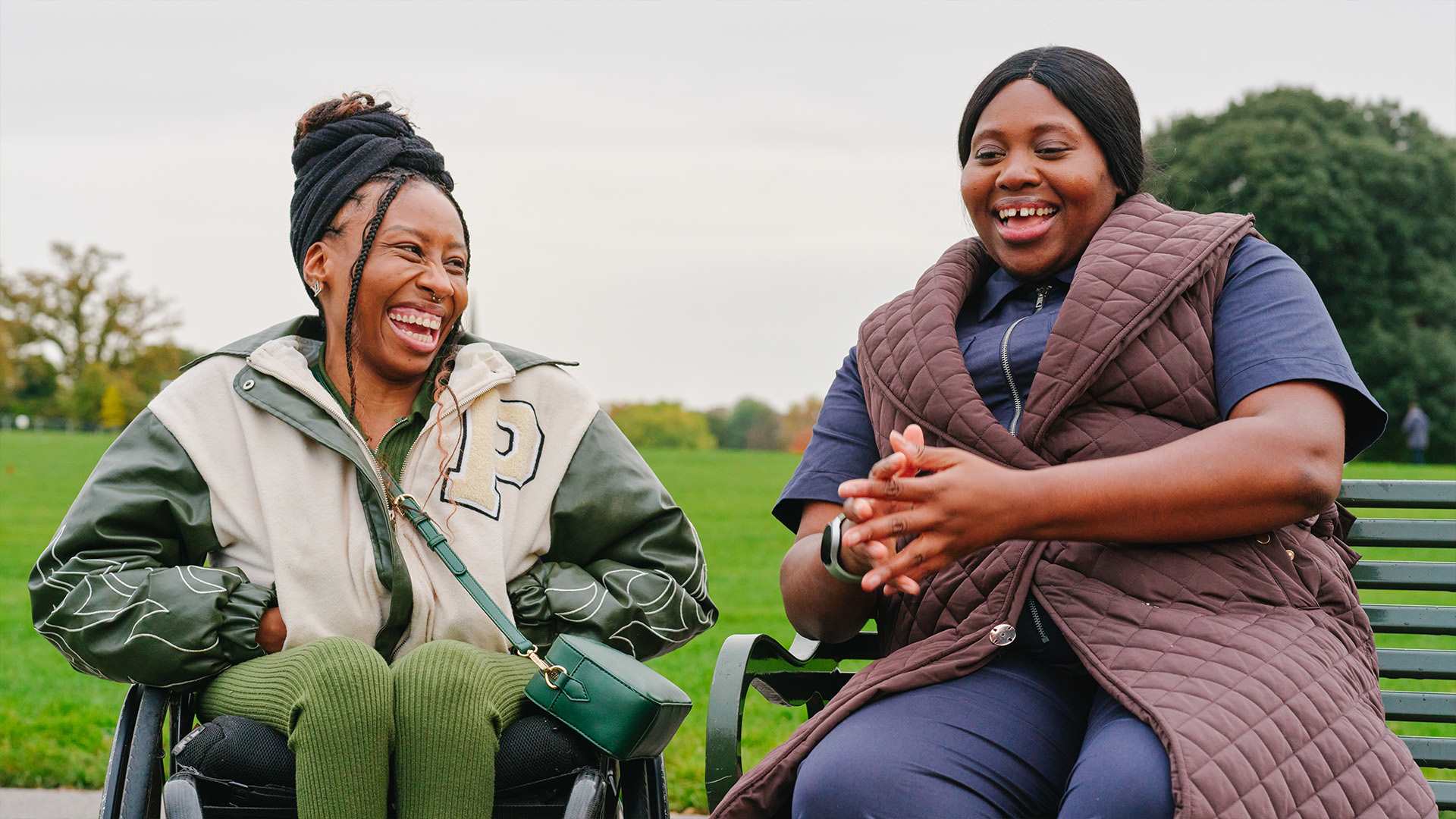 A young Black woman in a wheelchair and an older Black woman sitting on a bench in the park. They are laughing together.