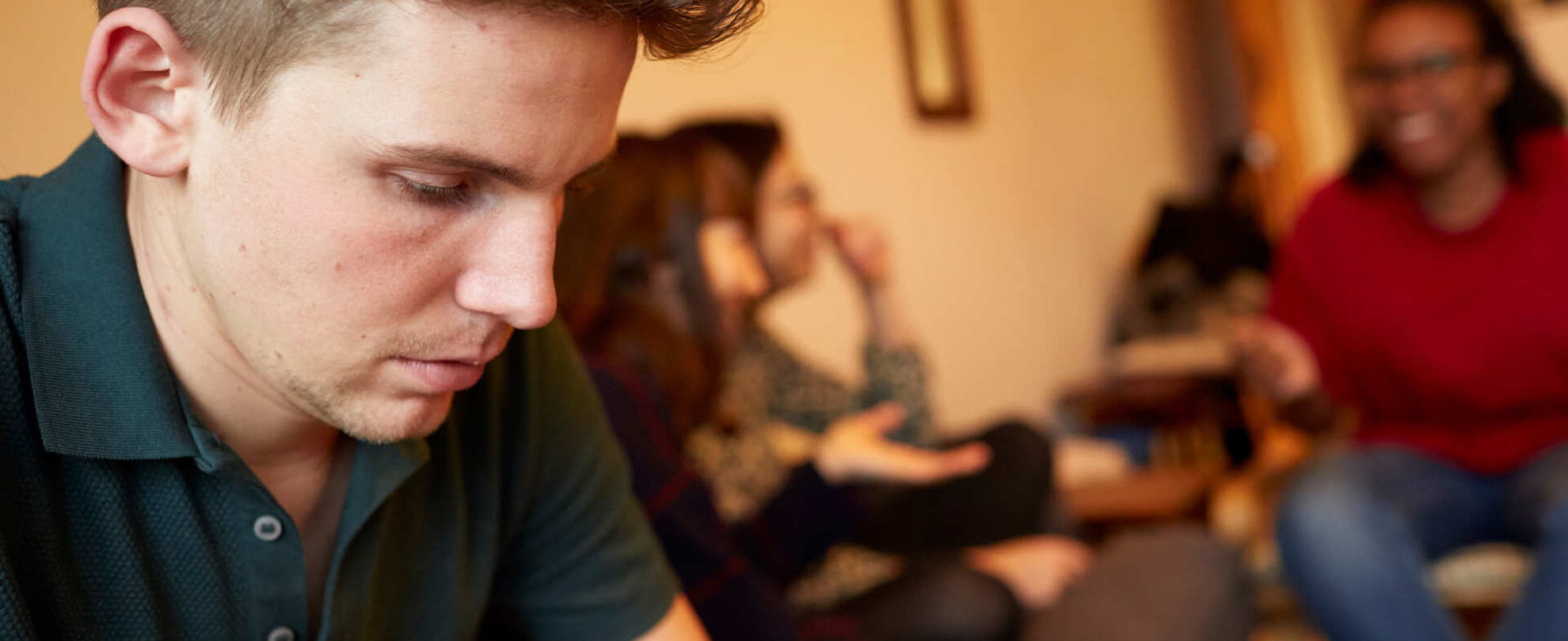 A young man looks worried and alone, while his three friends are talking and laughing in the background.