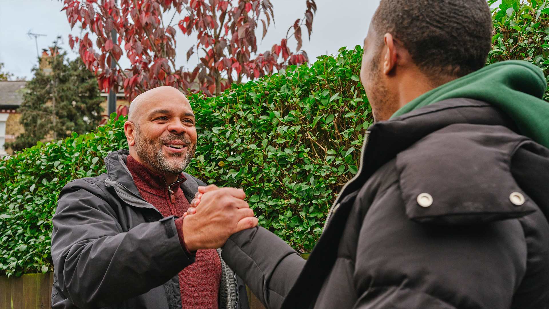 A young Black man grasping hands with an older Black man on the street. They are both smiling.