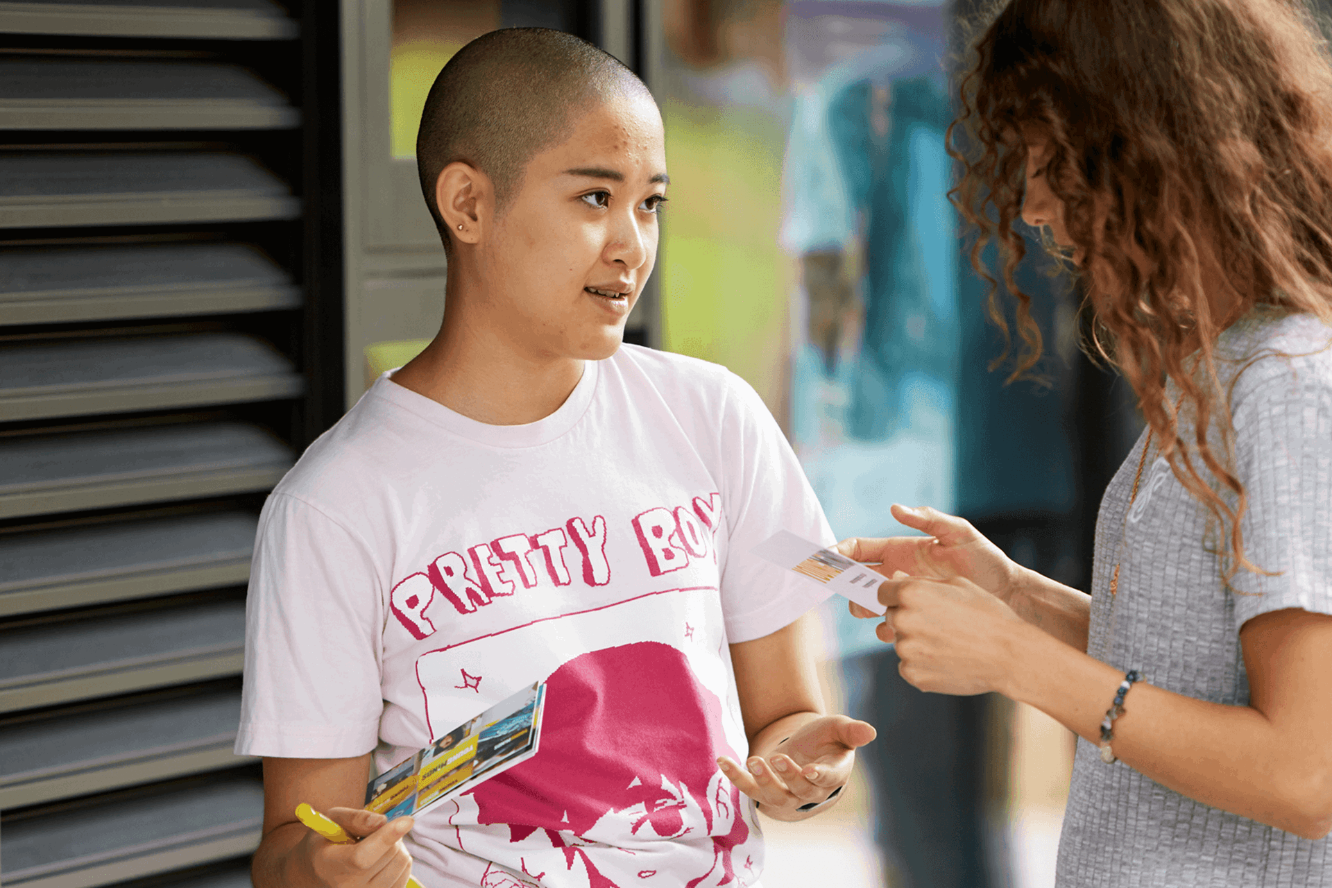 medium-shot-of-a-young-woman-with-shaved-head-holding-some-flyers-talking-to-another-girl-with-curly-hair-and-face-unseen-inside-campus