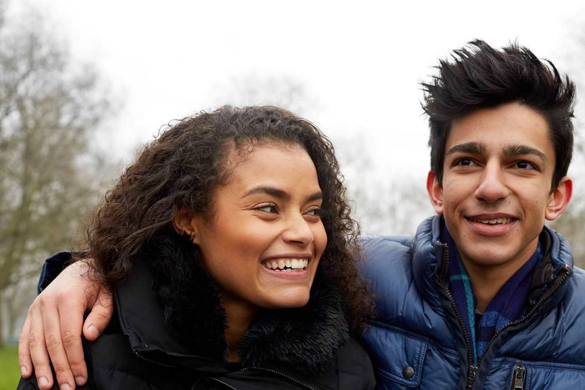 Two young people standing together and smiling, one has their arm around the shoulder of the other.