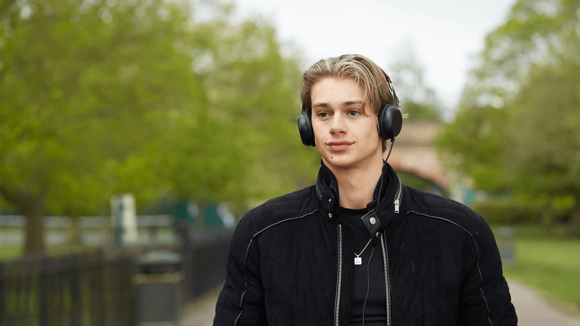 a boy wearing black jacket and with headphones on while walking on a tree lined street