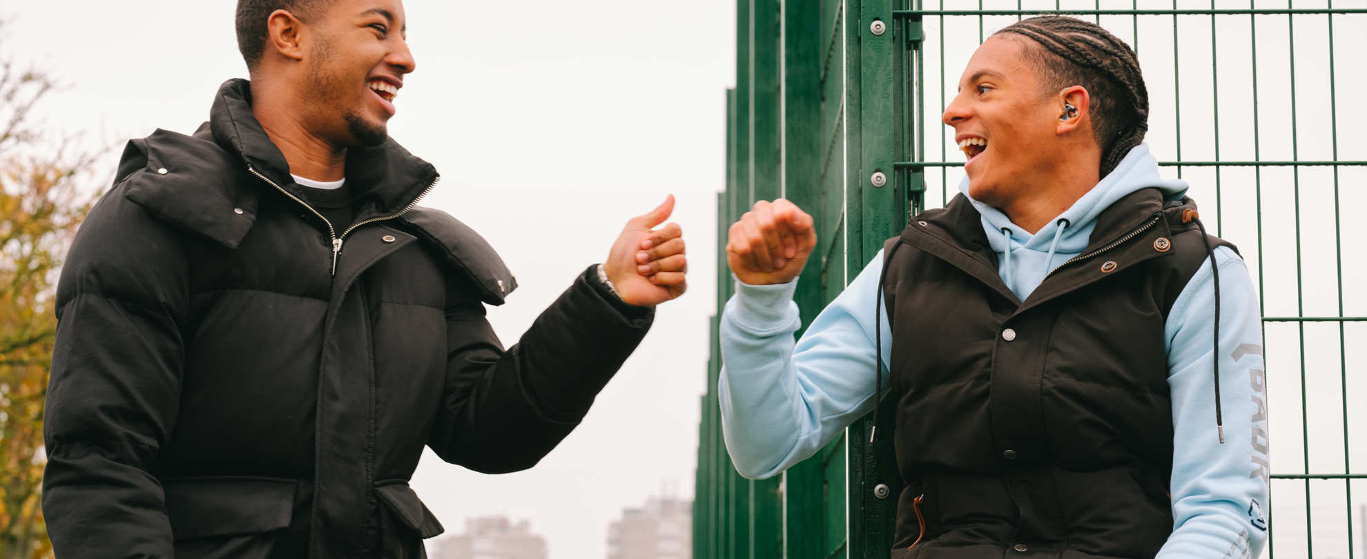 A Black teenage boy wearing a hearing aid bumping fists with a young Black man in the park.