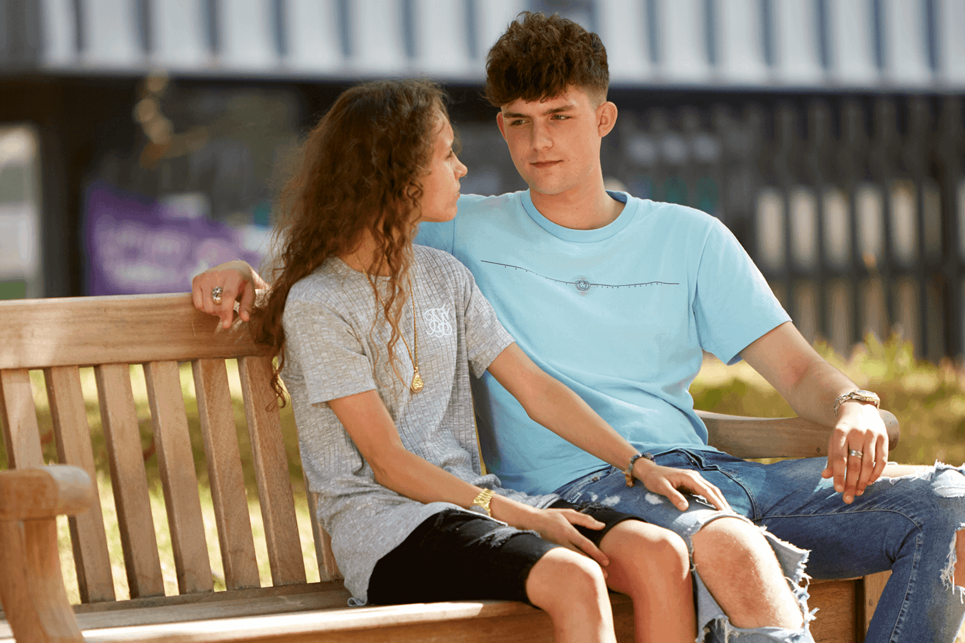 wide-shot-of-a-girl-and-a-boy-sitting-on-a-bench-while-looking-at-each-other-with-the-boy-arm-wrapped-on-her-shoulder-and-school-campus-on-background