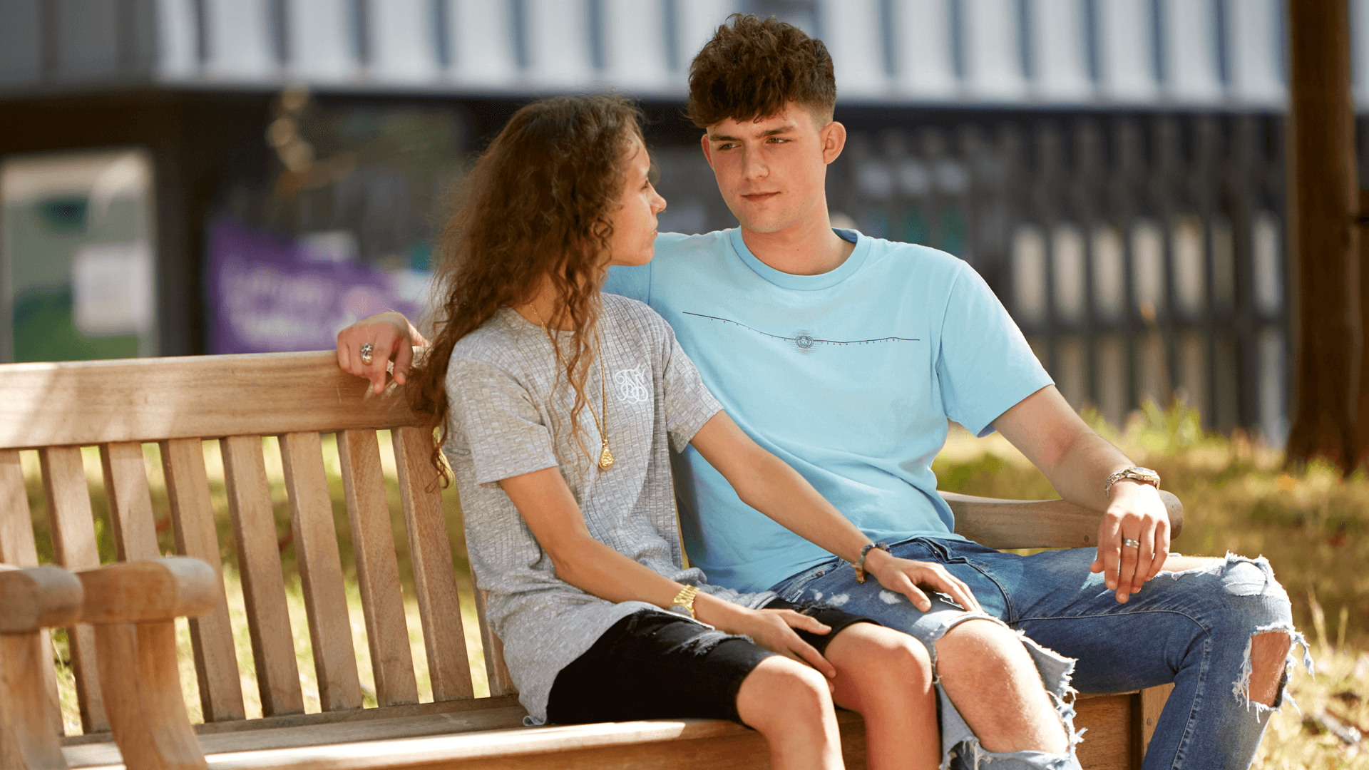 wide-shot-of-a-girl-and-a-boy-sitting-on-a-bench-while-looking-at-each-other-with-the-boy-arm-wrapped-on-her-shoulder-and-school-campus-on-background