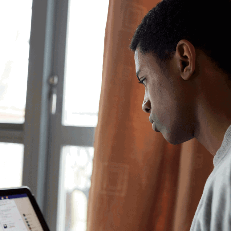 A boy wearing a grey t-shirt sits beside a window while using Facebook on his laptop.
