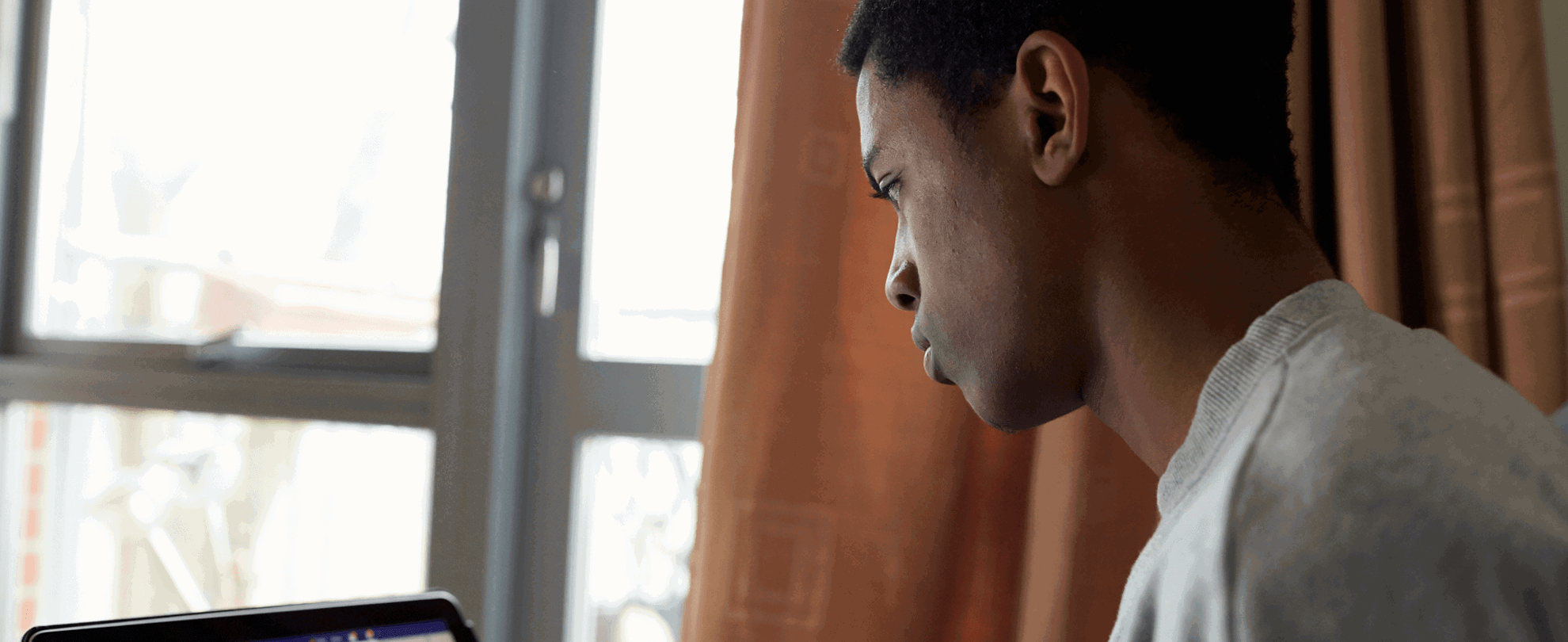 A boy wearing a grey t-shirt sits beside a window while using Facebook on his laptop.