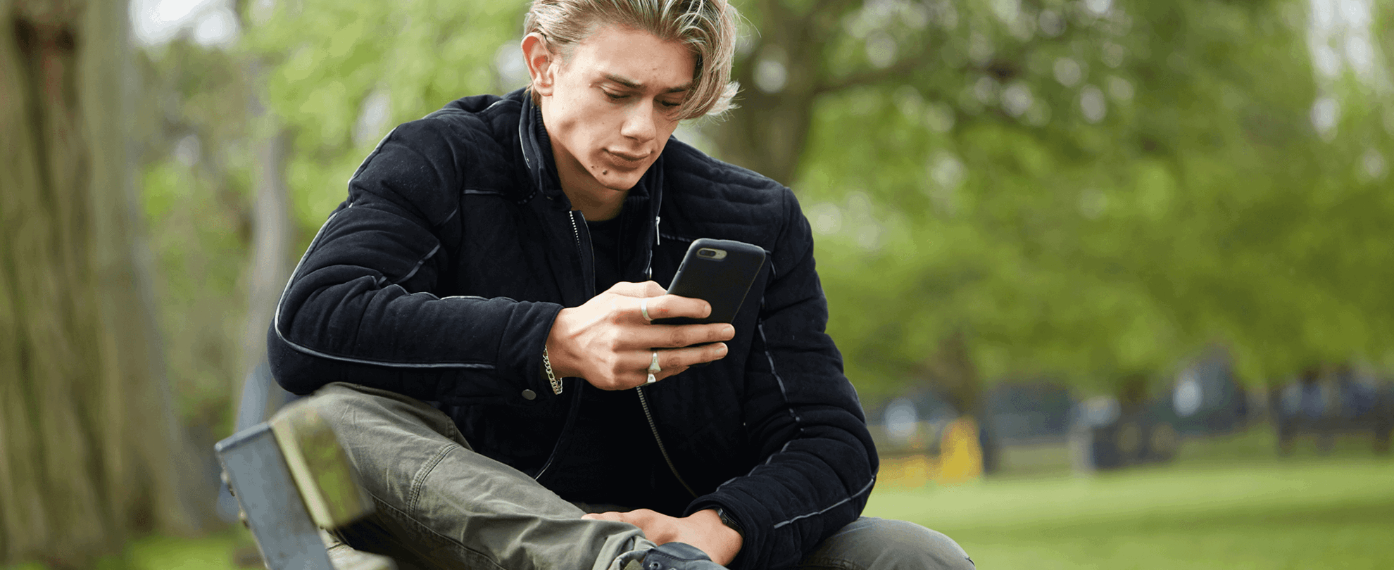 A young man wearing a black jacket sits on a park bench. He is looking at his phone with a worried expression.