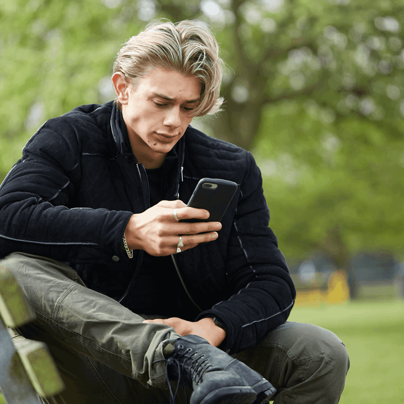 A young man wearing a black jacket sits on a park bench. He is looking at his phone with a worried expression.