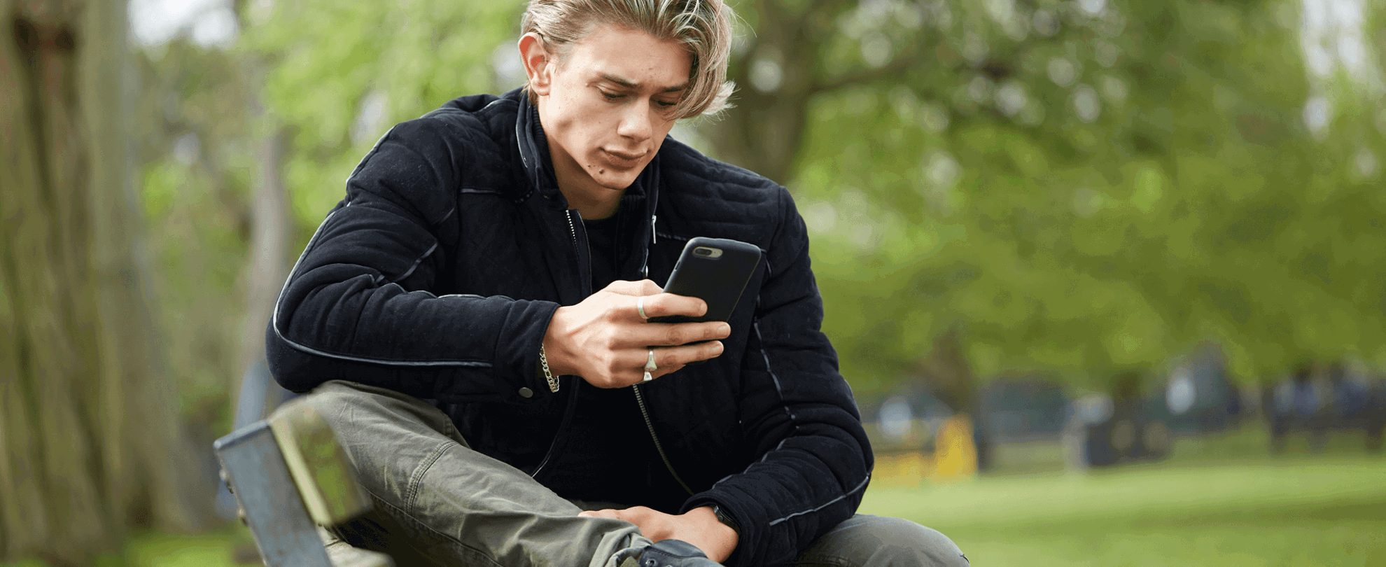 A young man wearing a black jacket sits on a park bench. He is looking at his phone with a worried expression.