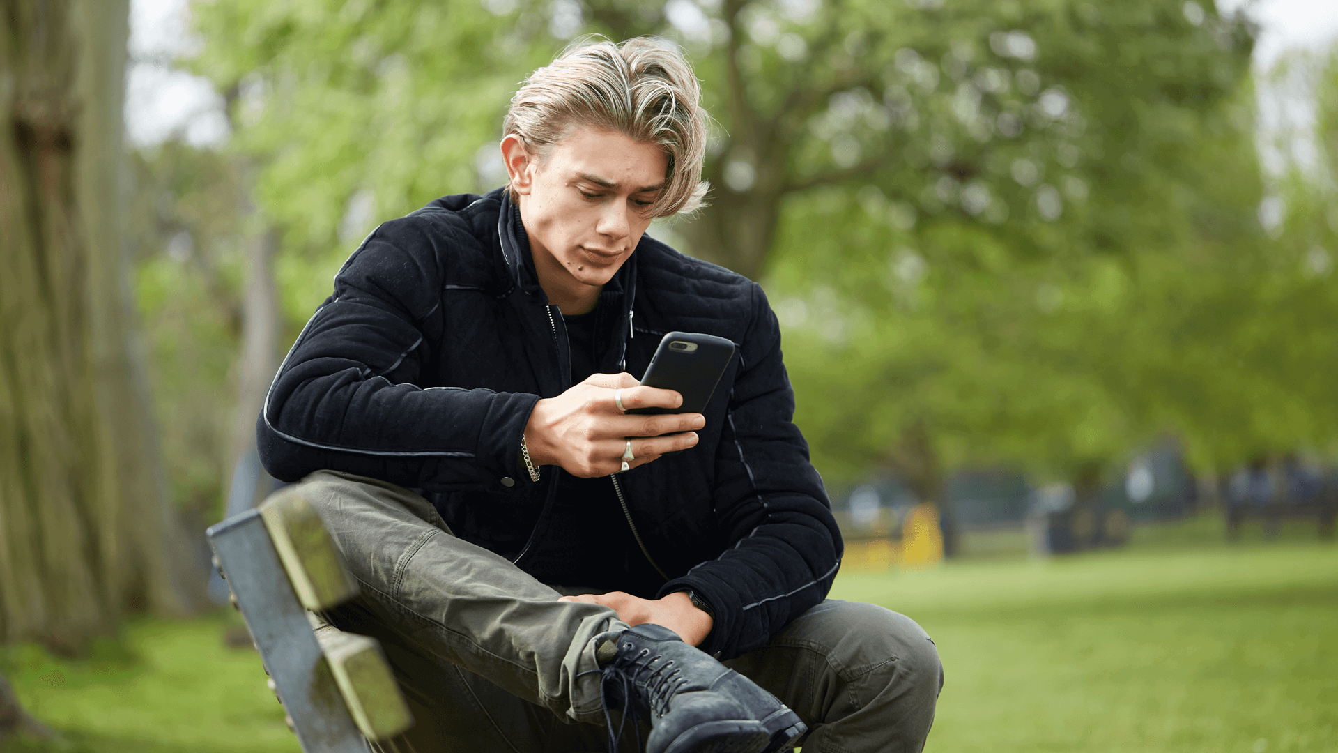 A young man wearing a black jacket sits on a park bench. He is looking at his phone with a worried expression.