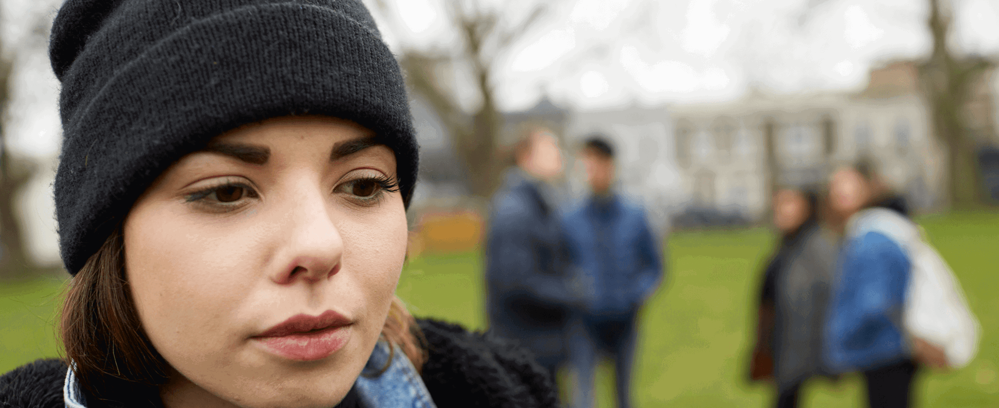 close up of a girl wearing a black beanie feeling left out by friends standing behind her with school and trees on background