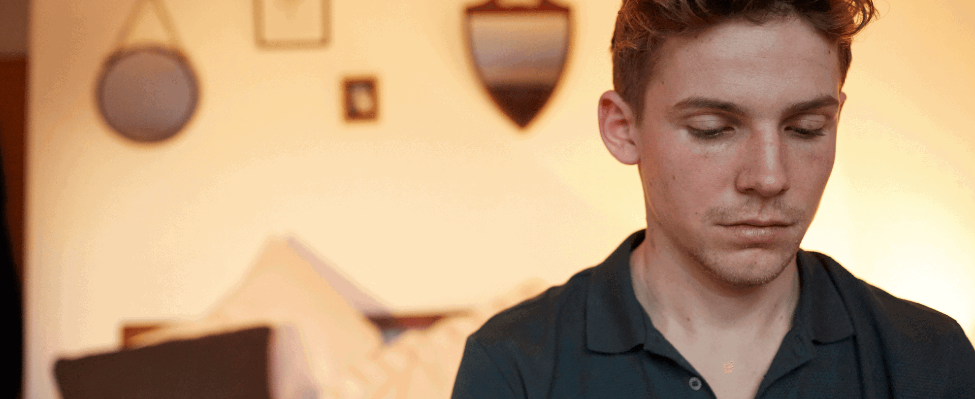 A boy sitting alone in his bedroom. He looks sad with his head and eyes facing down.