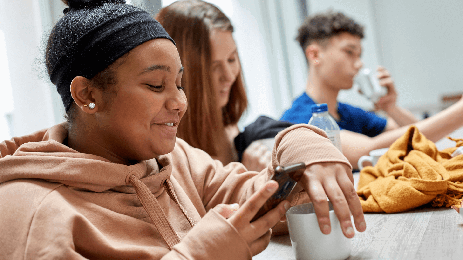 two girls smiling and looking at their phone while sitting on a dining bench having their lunch and a boy drinking water on the background