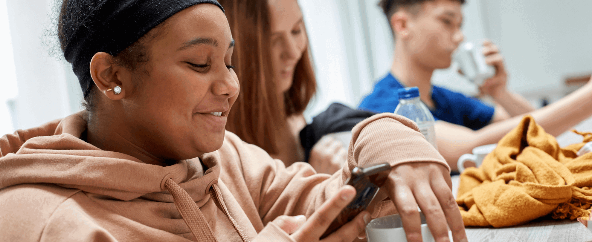 two girls smiling and looking at their phone while sitting on a dining bench having their lunch and a boy drinking water on the background