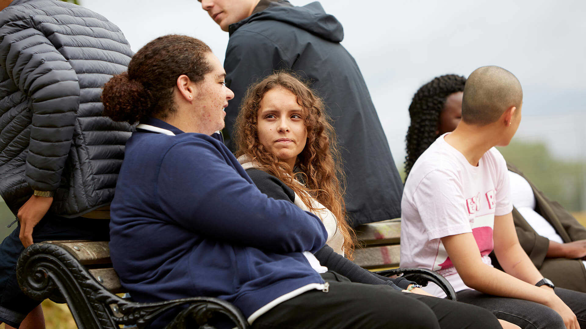 A group of six young people sitting on a bench and talking to each other in a park.