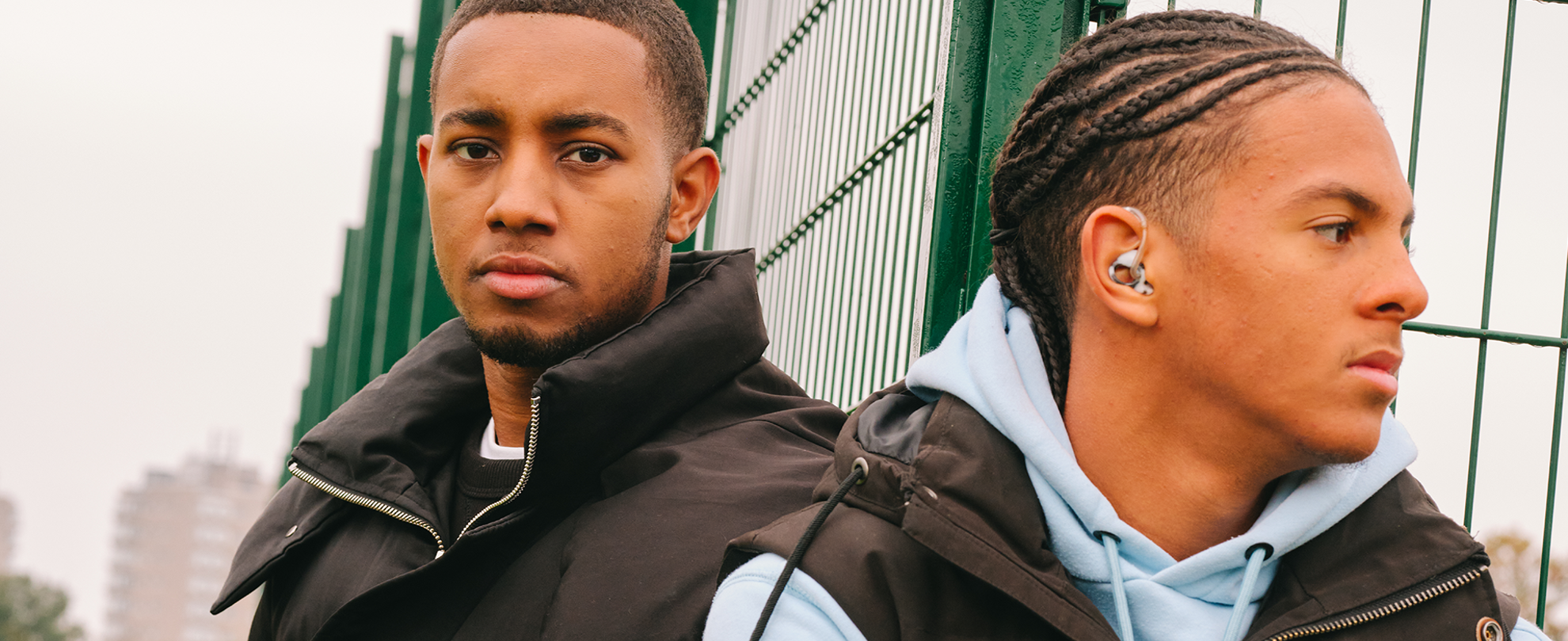 A young Black man sitting in the park with a Black teenage boy wearing a hearing aid. They are both looking very serious.