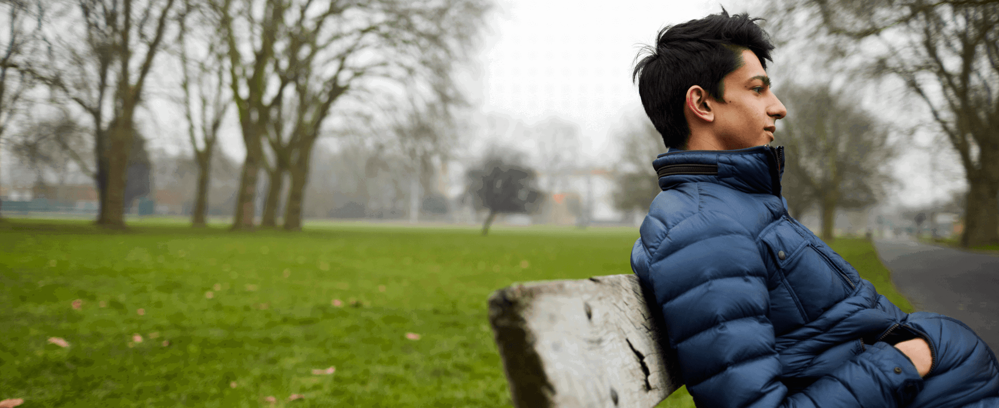 A boy sits on a park bench wearing a blue puffer jacket with his hands in his pockets. He is looking straight ahead as if deep in thought.