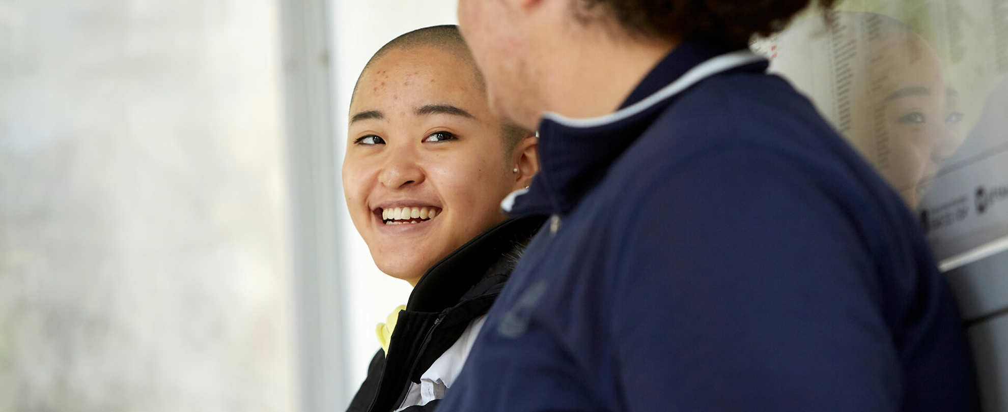 Two young people lean against a bus stop. The person on the left is laughing and wears a black coat while looking at the other young person. The person on the right wears a dark blue jacket and is looking at the person on the left. They're side on to the camera.
