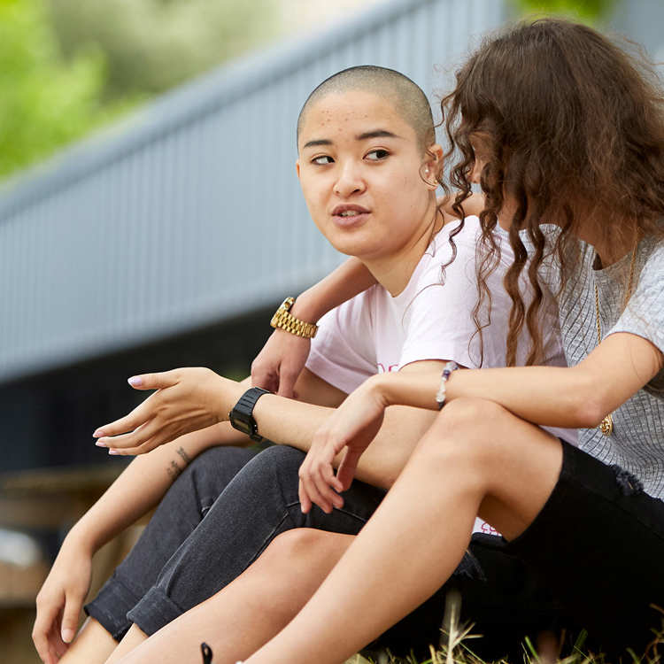 a-young-woman-with-curly-hair-and-in-grey-shirt-and-black-shorts-with-face-unseen-has-her-hands-wrapped-another-young-woman-with-shaved-hair-and-in-white-shirt-and-blakc-trousers-talking-to-her-while-they-sat-on-the-grass-with-a-tree-and-building-on-their-background
