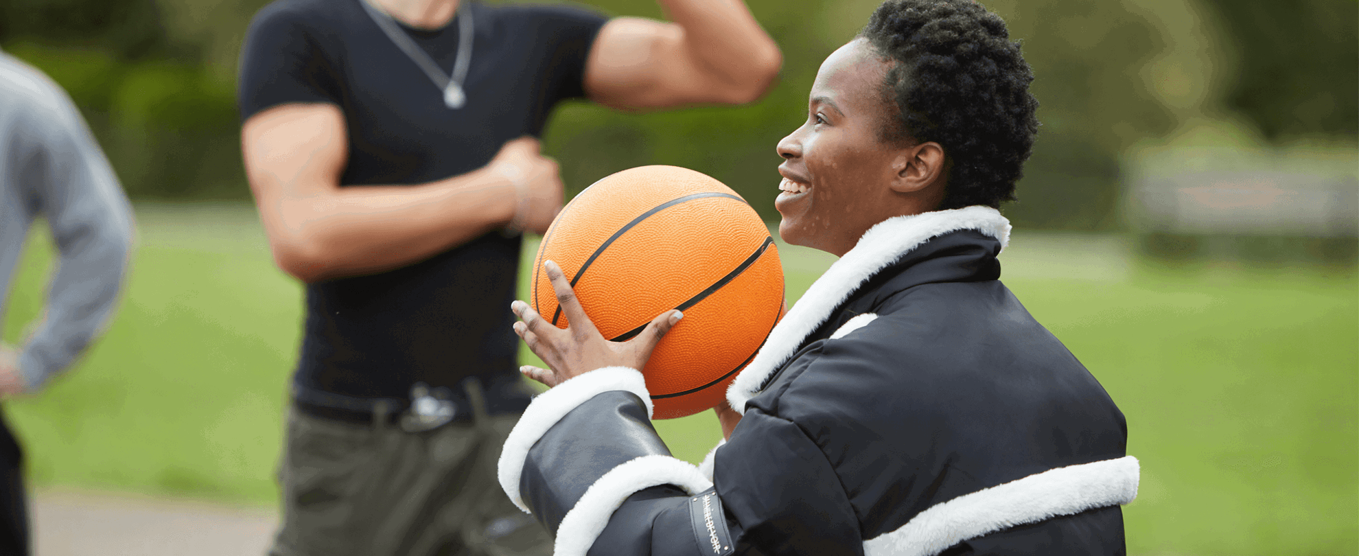 medium shot of a girl with short curly hair wearing black jacket and holding a basketball while smiling and playing with boys