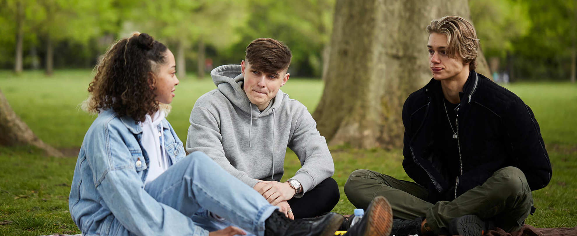 wide-shot-of-three-young-people-sitting-on-the-ground-together-talking-to-each-other-and-listening-with-trees-on-their-background