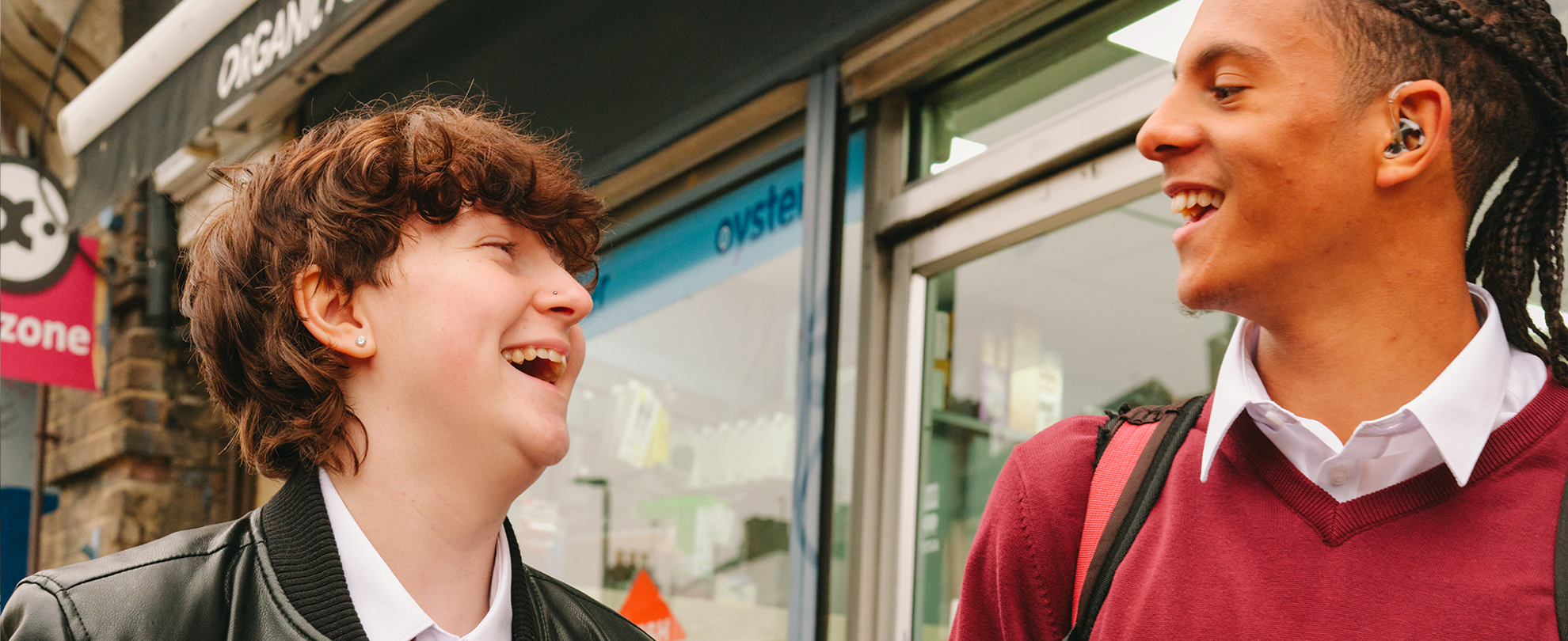 A Black teenage boy wearing a hearing aid laughing with a white non-binary teenager outside the shops.