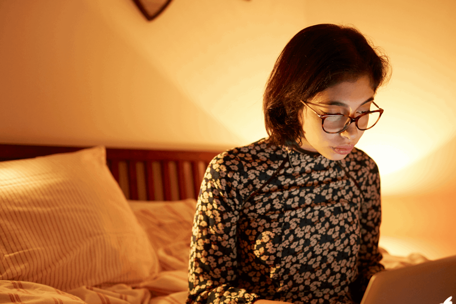 A girl wearing glasses sitting on the end of her bed while using her laptop.