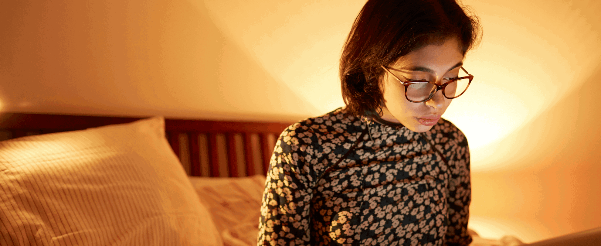A girl wearing glasses sitting on the end of her bed while using her laptop.