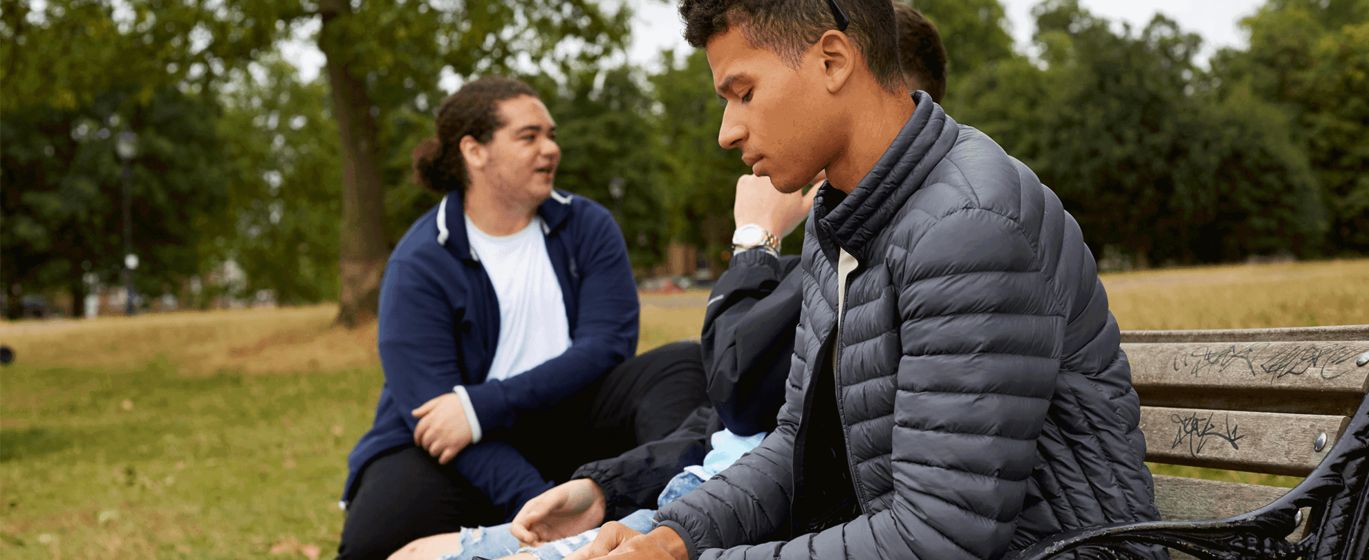 boy-in-a-jacket-looking-at-his-phone-ignoring-his-friends-who-are-talking-beside-him-while-sitting-on-a-bench-at-the-park
