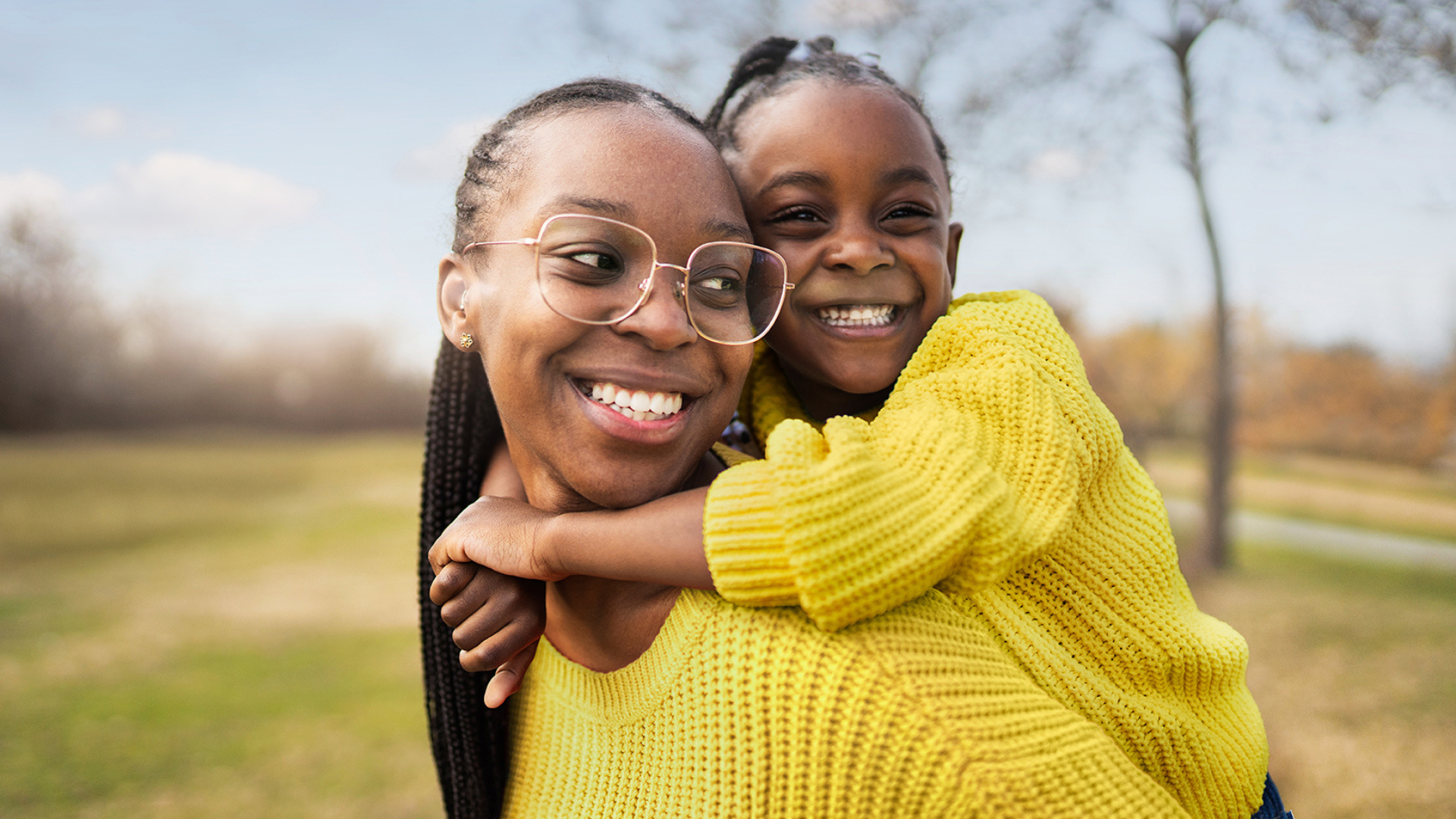 A mother with a young daughter on her back, both smiling and in bright yellow jumpers.