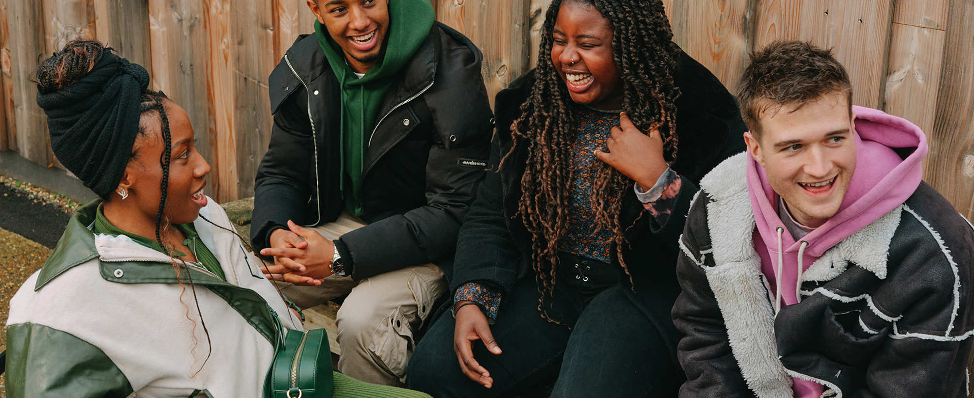 A group of young people laughing together outside on a bench. Group includes two Black young women (one in a wheelchair), one Black young man, and a white young man.