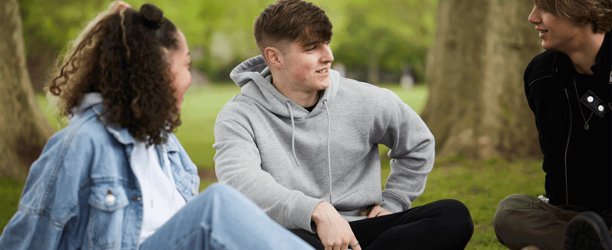 Three young people talking while sitting in the grass in a park.