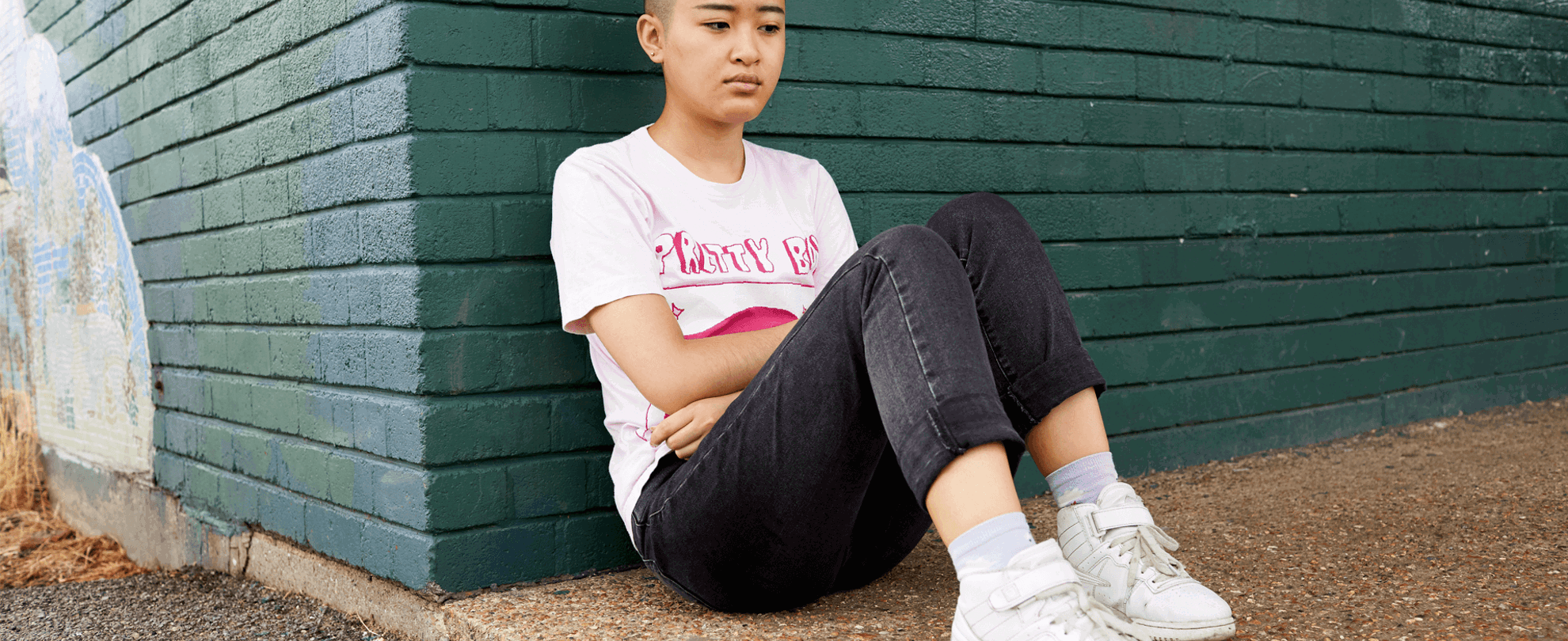 A girl with a shaved head wearing a t-shirt and black jeans. She is sitting on the ground and leaning against a green brick wall.