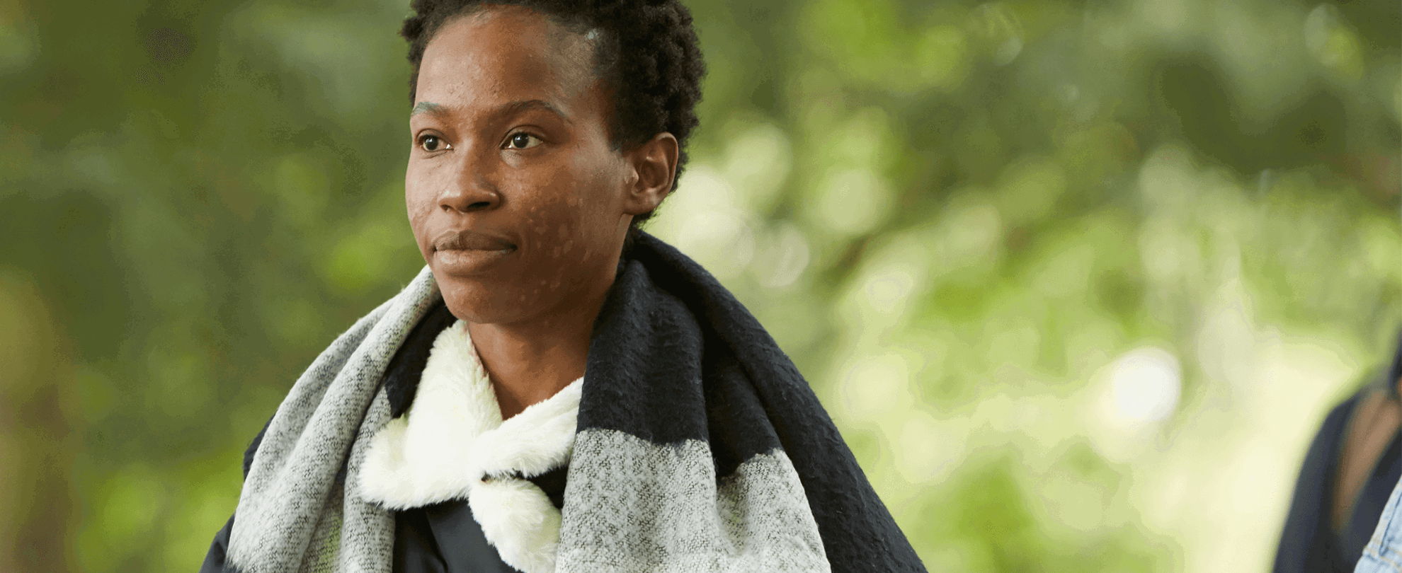 A young woman wearing a grey and black scarf walking through a park.