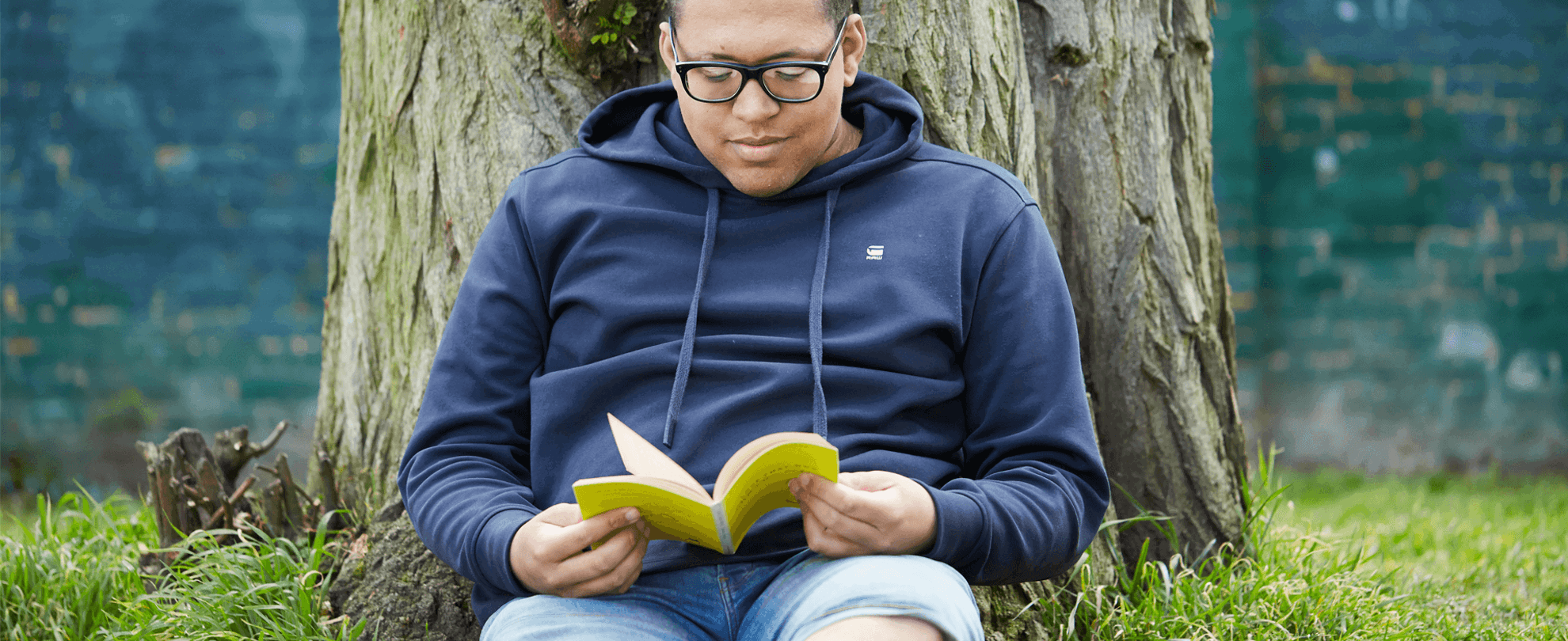 A young man wearing glasses and a navy hoodie. He is sat on the grass and leaning against a tree while reading a book.