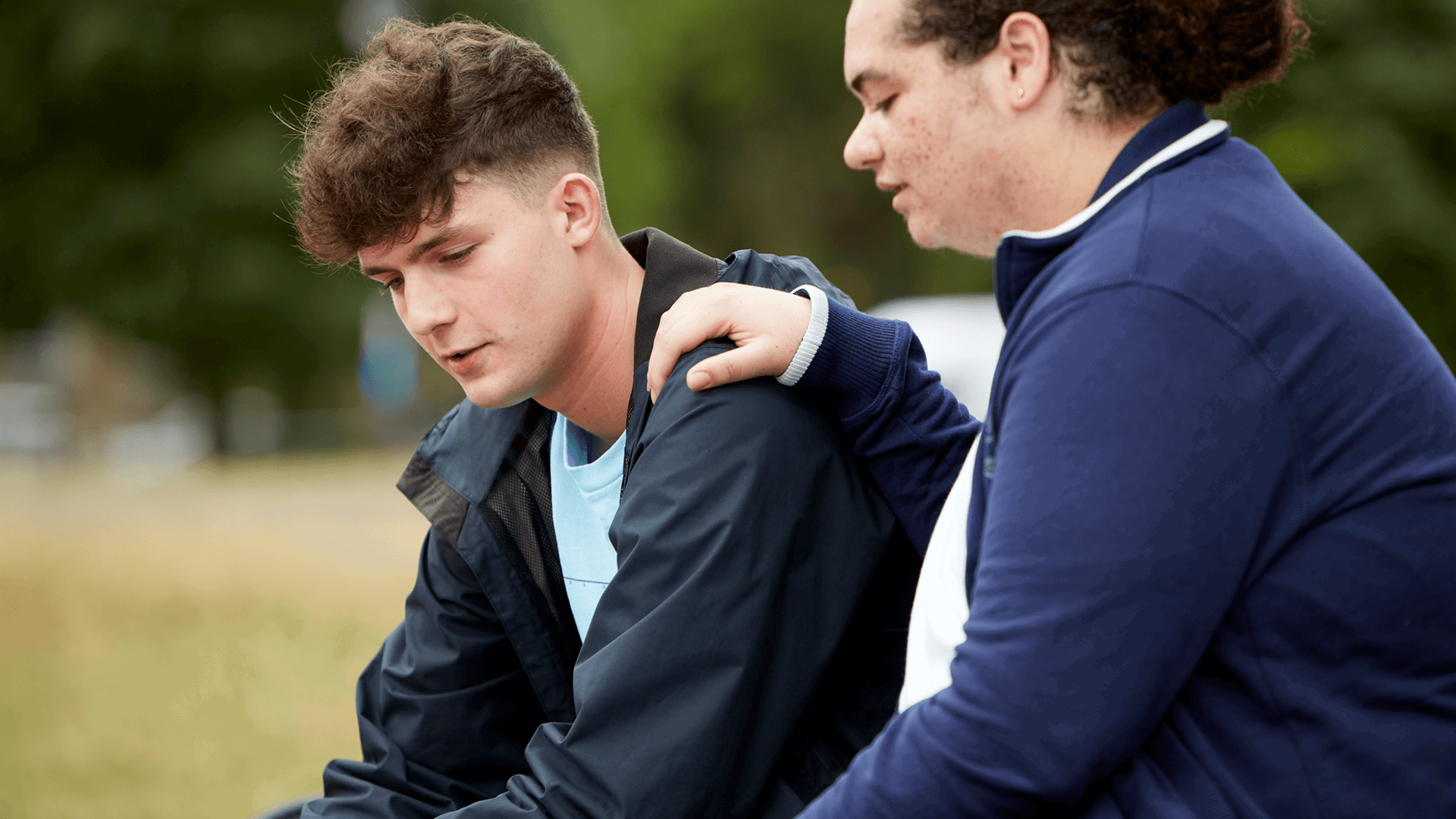 A boy comforting his upset friend by putting with his hand on his shoulder while they sit in a park.