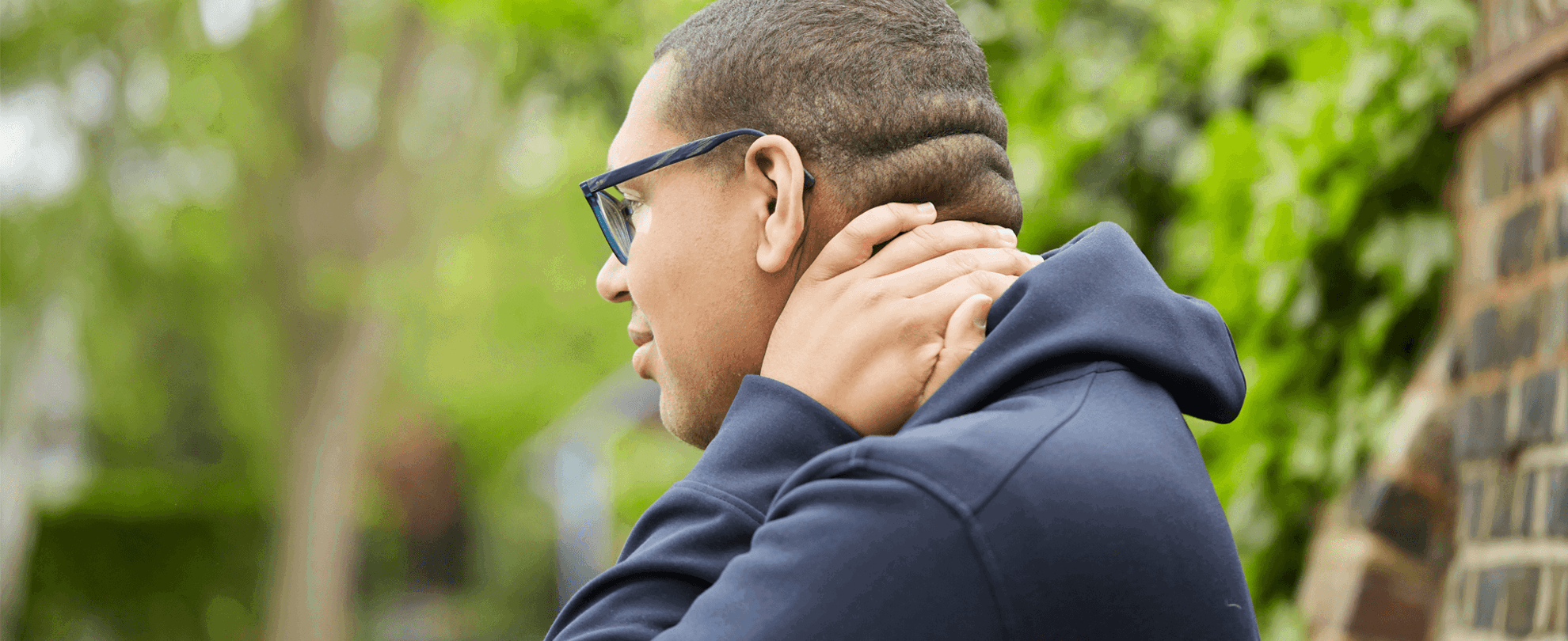 A boy wearing glasses and a black hoodie stands in a park looking worried. He is rubbing the back of his neck with one hand.