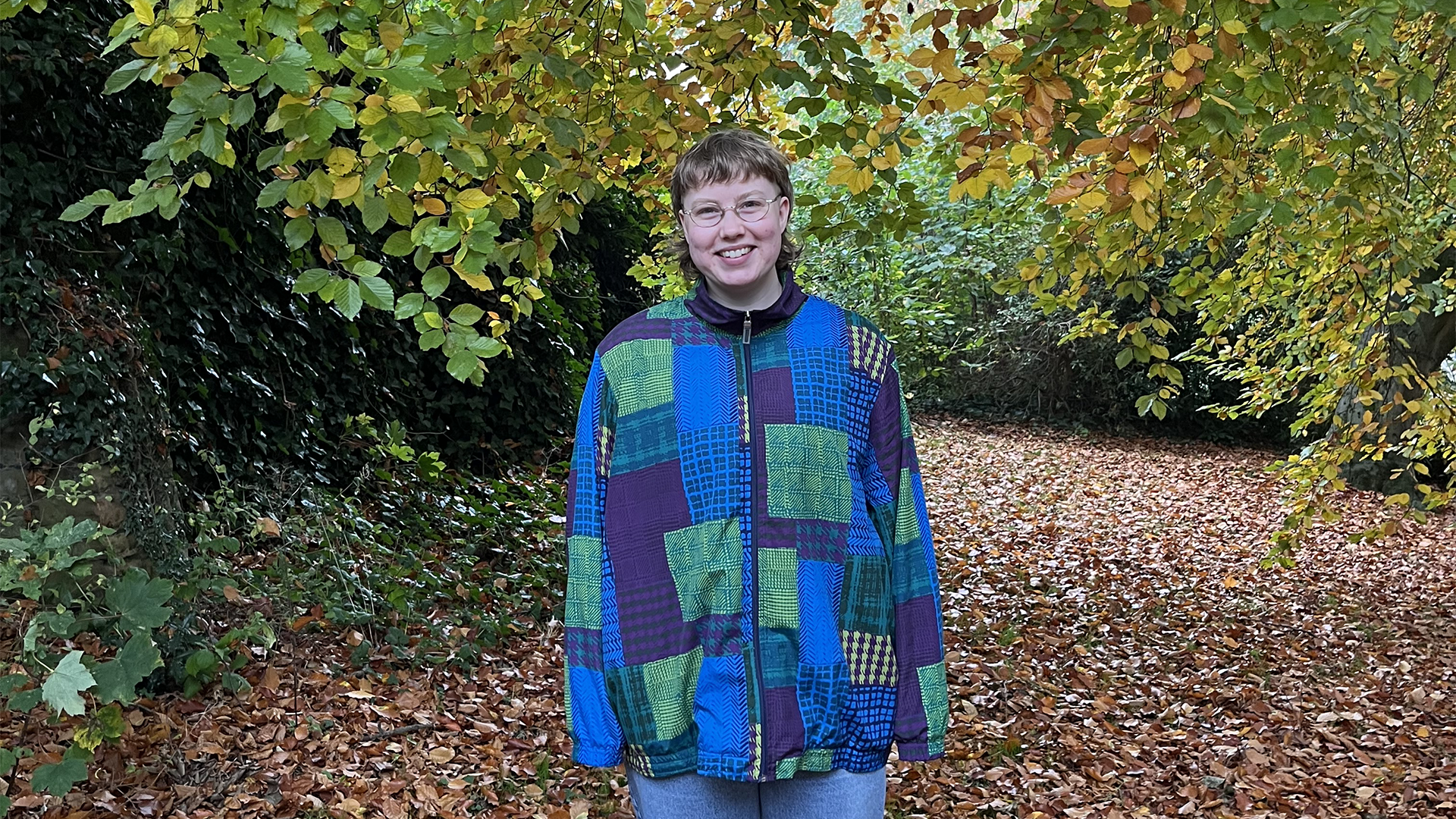Sofe standing in a forest and smiling. They are wearing glasses and a blue and green patterned jacket.