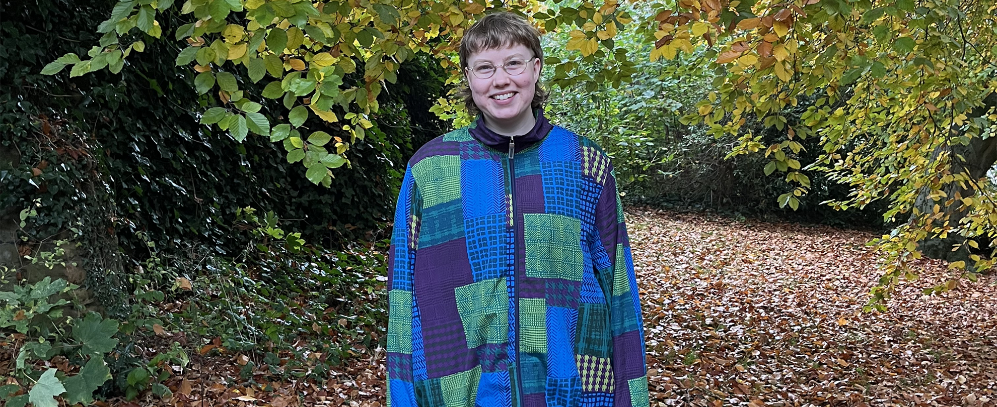 Sofe standing in a forest and smiling. They are wearing glasses and a blue and green patterned jacket.