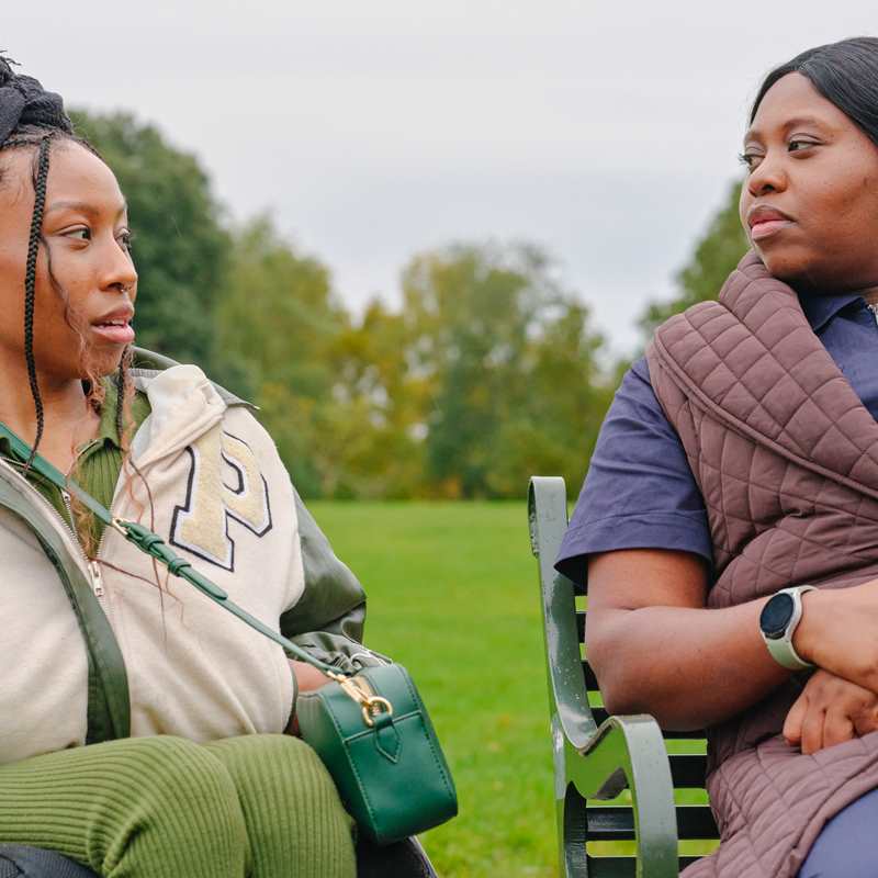 A young Black woman in a wheelchair talking to an older Black woman on a bench in the park.