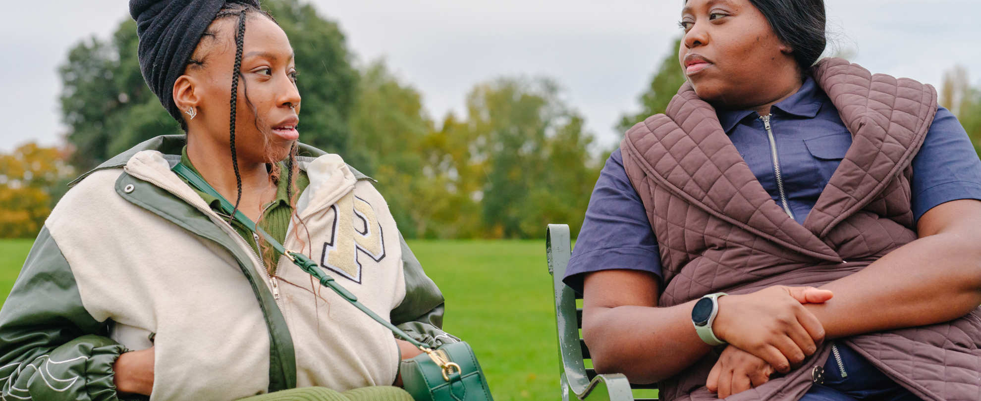 A young Black woman in a wheelchair talking to an older Black woman on a bench in the park.
