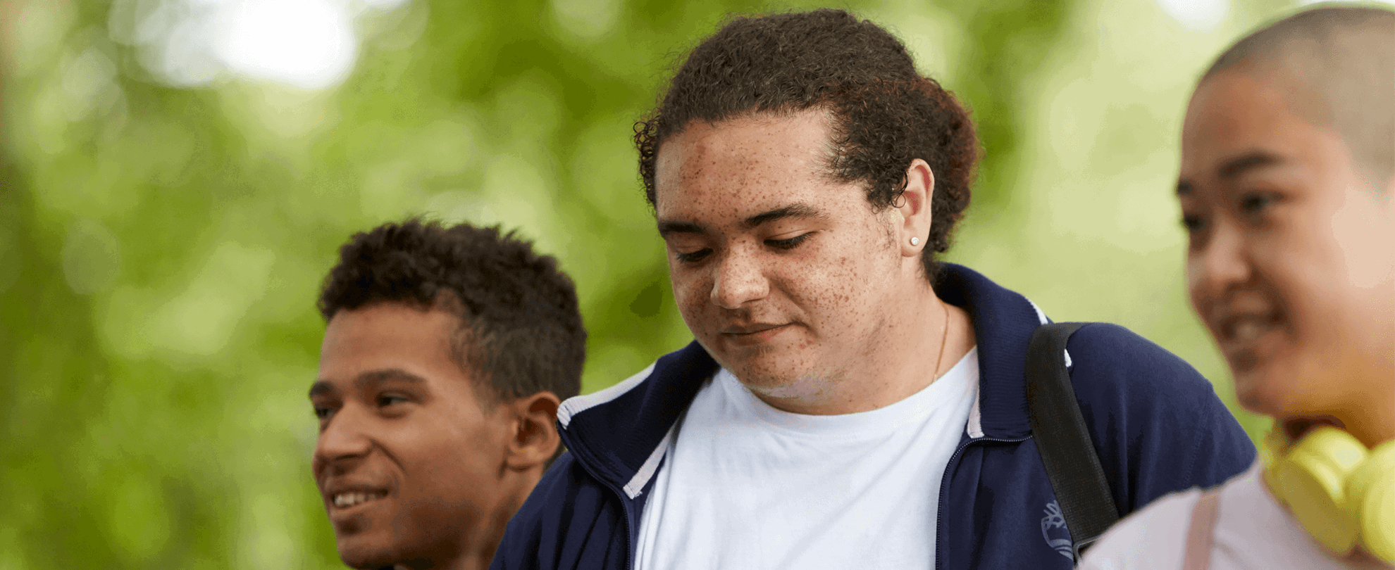 Three young people walking together through a park. Two of them are smiling while a boy in the centre looks worried.